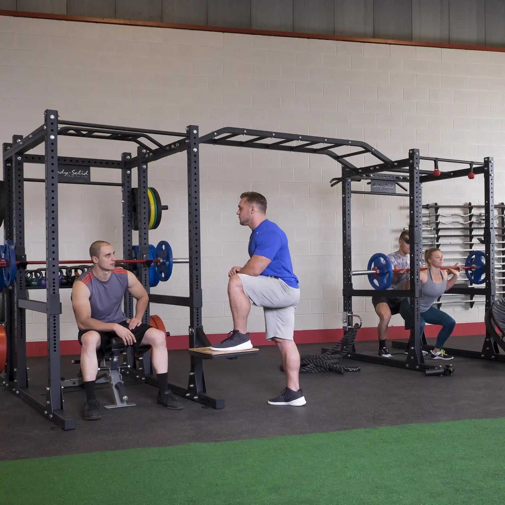 Gym setting with people using the Body-Solid Pro ClubLine Double Extended Commercial Power Rack SPR1000DBBACK for various exercises, including squats and bench presses.