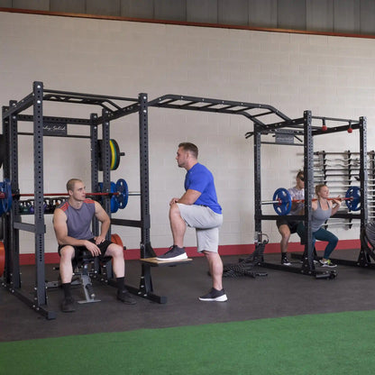 Gym setting with people using the Body-Solid Pro ClubLine Double Extended Commercial Power Rack SPR1000DBBACK for various exercises, including squats and bench presses.