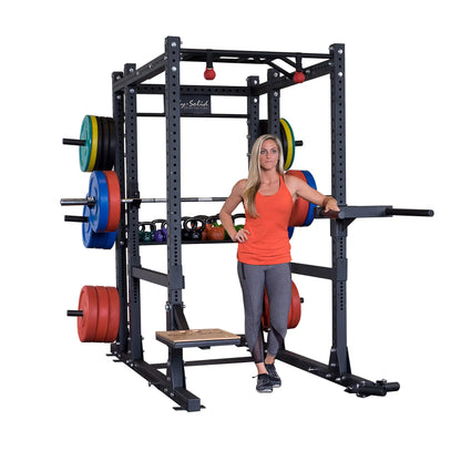 A woman in athletic clothing stands inside a Body-Solid Pro ClubLine Extended Commercial Power Rack SPR1000BACK, with various weights and kettlebells organized around the rack.