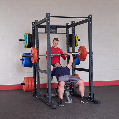 A person bench pressing on the Body-Solid Pro ClubLine Extended Commercial Power Rack SPR1000BACK, assisted by a spotter, with colorful weight plates on either side.