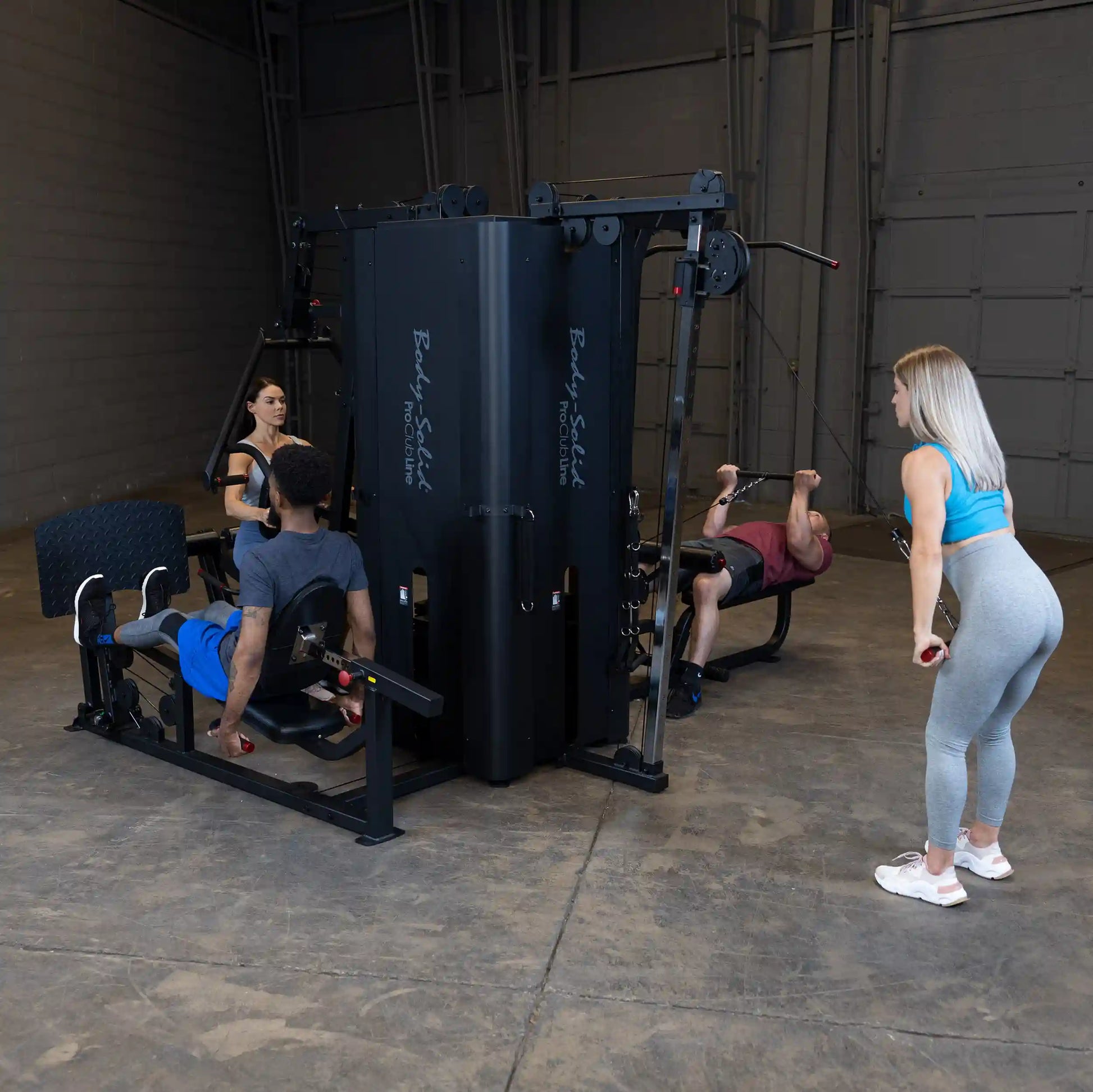 A fitness scene featuring the Body-Solid Pro ClubLine Four-Stack Gym S1000, with individuals performing various exercises, including seated leg curls and cable exercises, in a spacious gym setting.