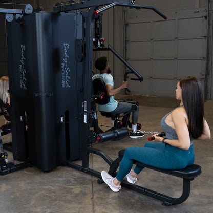 A fitness scene featuring the Body-Solid Pro ClubLine Four-Stack Gym S1000, with two individuals engaged in exercises; one person seated and using the machine's handles while the other performs a different workout on a connected station.