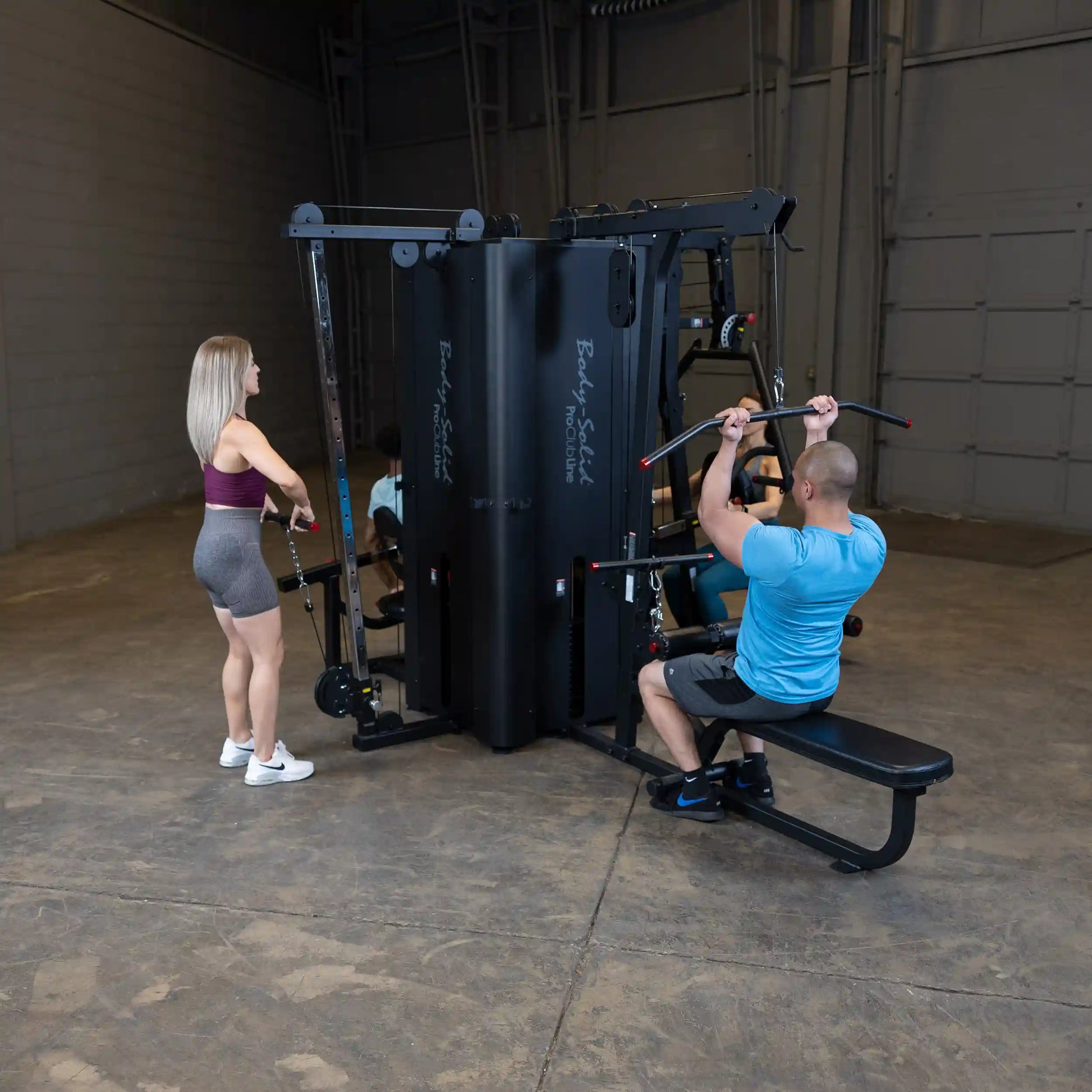 A gym setting featuring the Body-Solid Pro ClubLine Four-Stack Gym S1000, with two individuals using the equipment for their workouts. One person is seated and performing a lat pulldown, while the other is standing and preparing for a cable exercise.