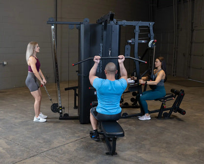 A fitness scene featuring the Body-Solid Pro ClubLine Four-Stack Gym S1000, with two women and one man exercising; one woman is using a cable machine while the man performs a pull-down exercise, and another woman is seated on a machine.