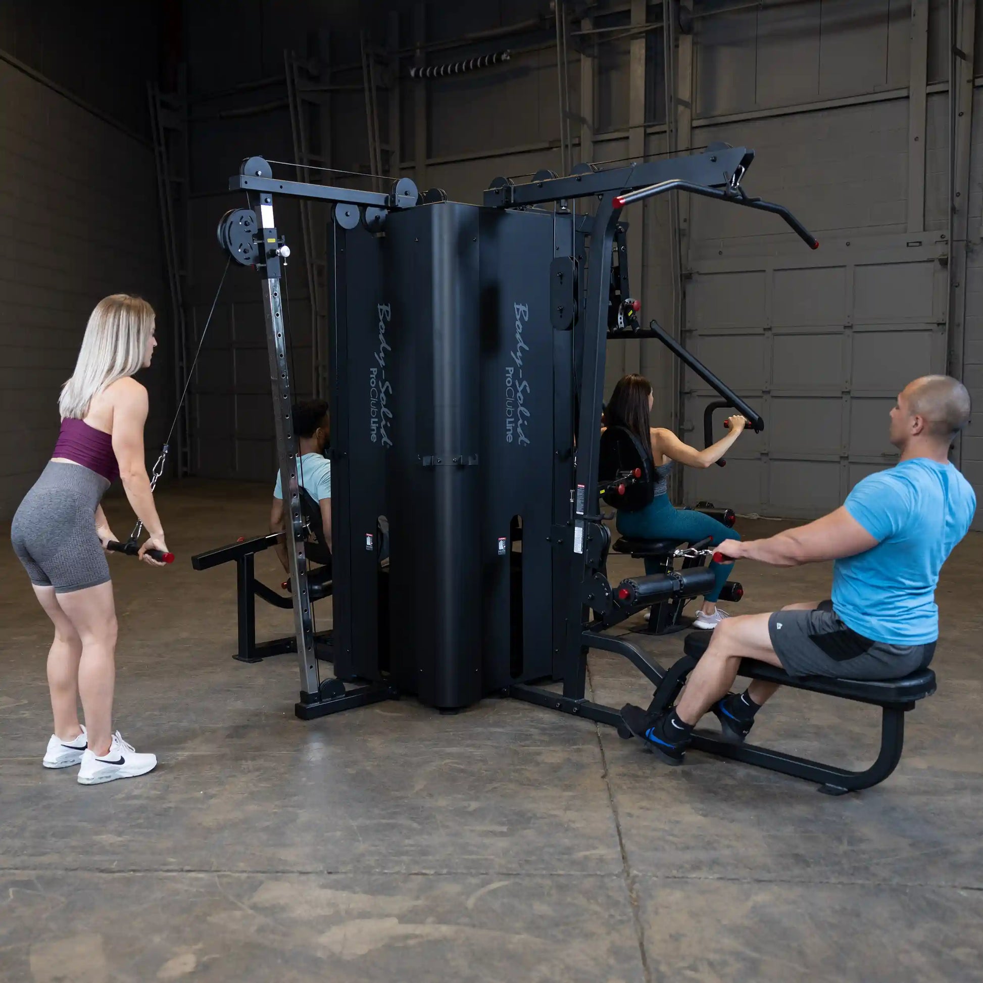 Three individuals exercising on the Body-Solid Pro ClubLine Four-Stack Gym S1000, with one person using the cable machine, another on a rowing machine, and a third on an adjacent station.