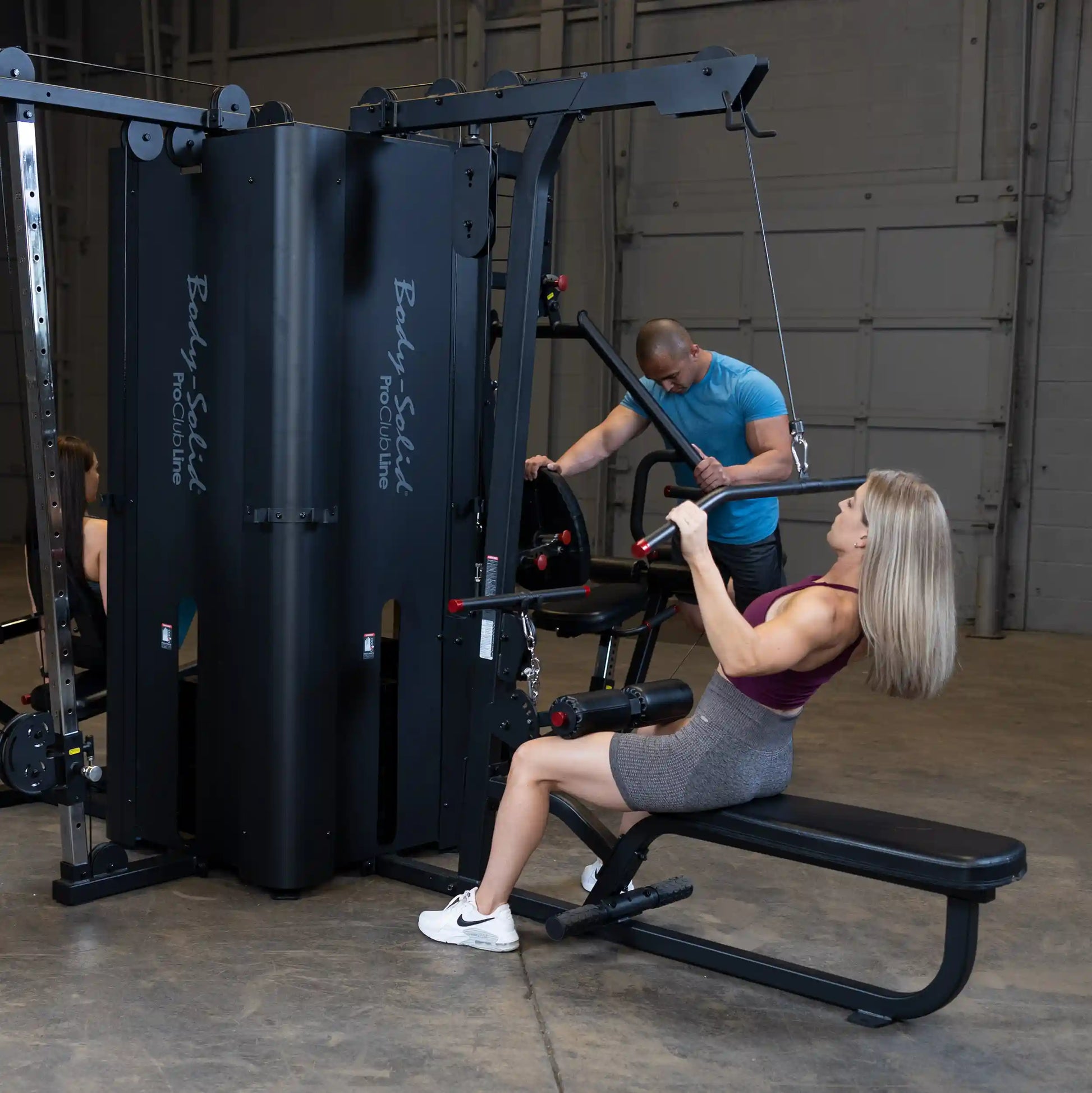 A woman using the Body-Solid Pro ClubLine Four-Stack Gym S1000 while a man adjusts equipment in the background, set in a gym environment.