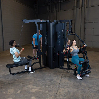 A fitness scene featuring the Body-Solid Pro ClubLine Four-Stack Gym S1000, with two individuals exercising on different machines, while another person observes in a spacious gym environment.