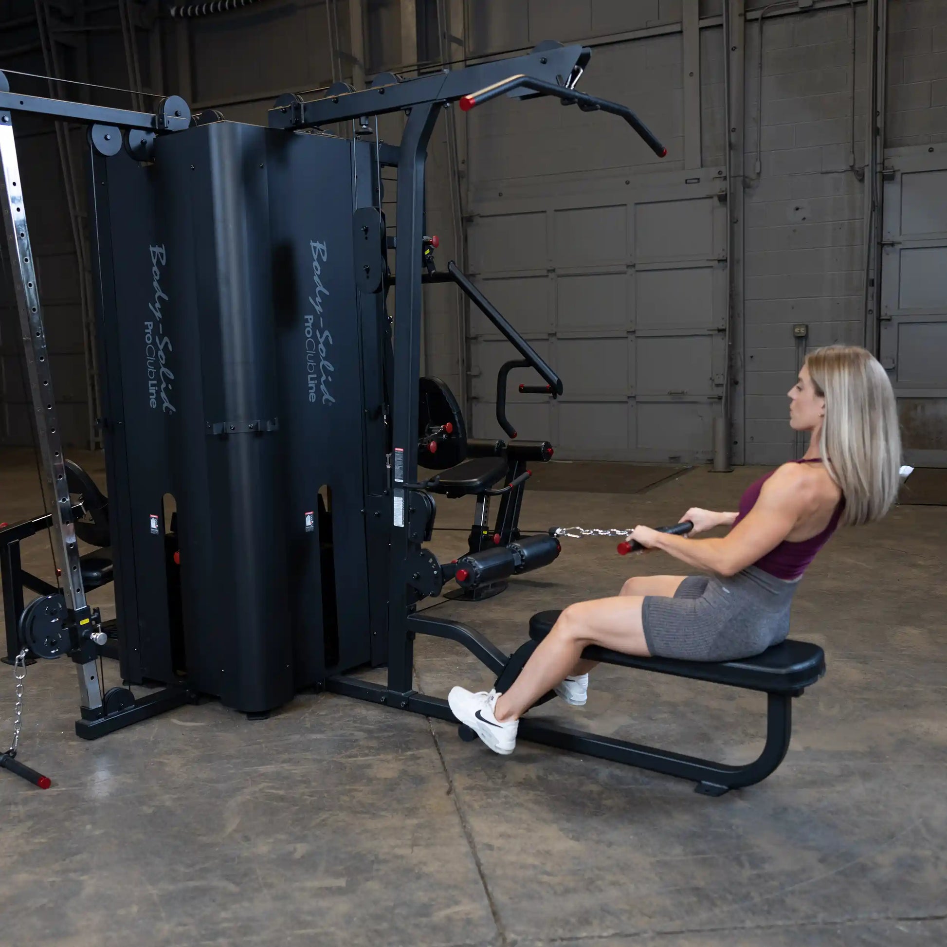 A woman using the Body-Solid Pro ClubLine Four-Stack Gym S1000 seated on a bench, performing a rowing exercise with a cable attachment in a spacious gym environment.