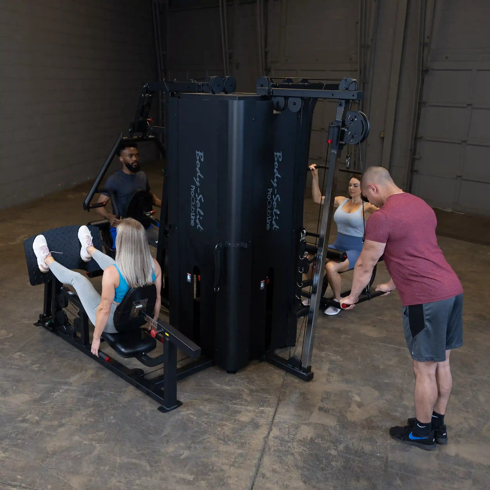 A fitness scene featuring the Body-Solid Pro ClubLine Four-Stack Gym S1000, with four individuals engaged in various exercises, including leg presses and cable pulls, in a spacious gym environment.