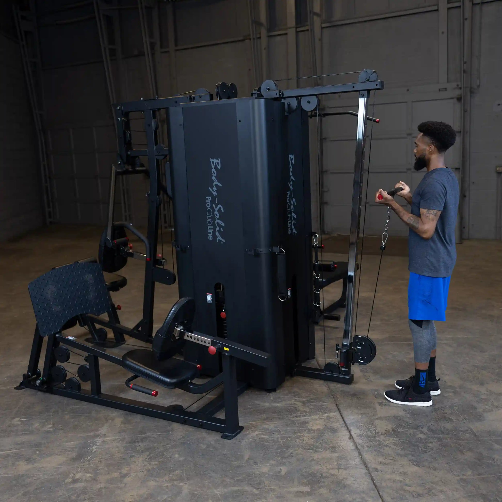 A person using the Body-Solid Pro ClubLine Four-Stack Gym S1000 in a spacious indoor gym setting, demonstrating exercise equipment with various attachments and a bench.