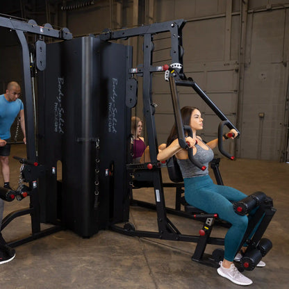 A fitness scene featuring the Body-Solid Pro ClubLine Four-Stack Gym S1000, with a woman performing an exercise on the machine while two other individuals observe in a gym setting.