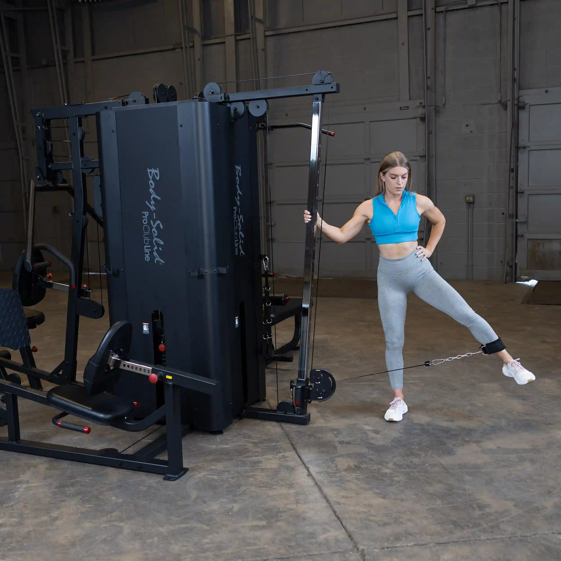 Woman exercising with the Body-Solid Pro ClubLine Four-Stack Gym S1000, performing a leg lift while using a cable attachment in a spacious gym setting.