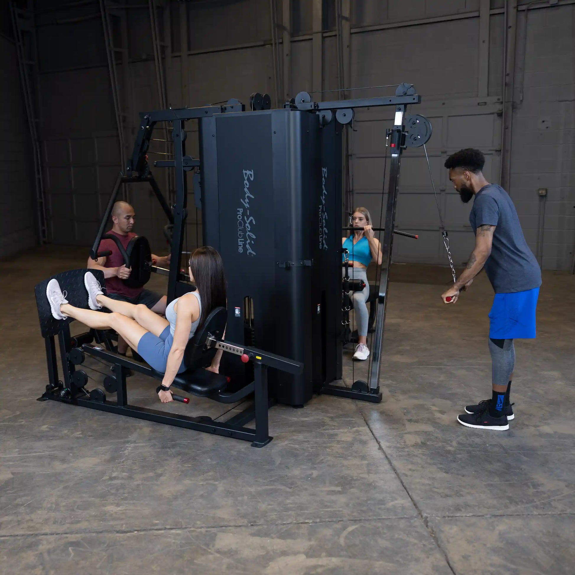 A group of four individuals using the Body-Solid Pro ClubLine Four-Stack Gym S1000 in a spacious gym setting. One person is performing leg presses, while others are engaged in various exercises on the multi-station machine.
