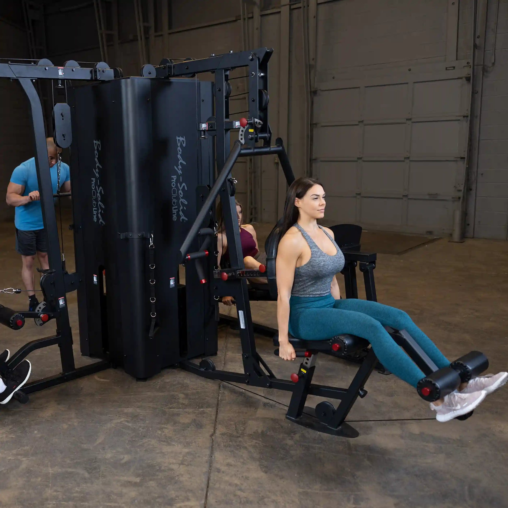 A woman using the Body-Solid Pro ClubLine Four-Stack Gym S1000 for leg exercises, with two other individuals in the background engaged in workouts in a gym setting.