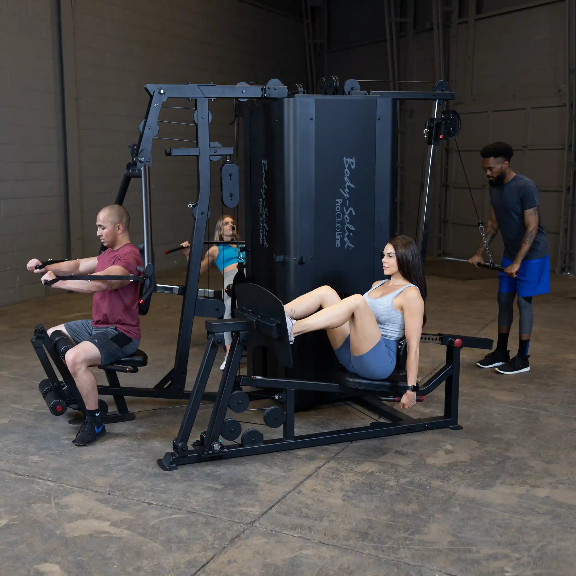 A fitness studio featuring the Body-Solid Pro ClubLine Four-Stack Gym S1000, with four individuals using different exercise stations, including a seated row, leg press, and cable machine.