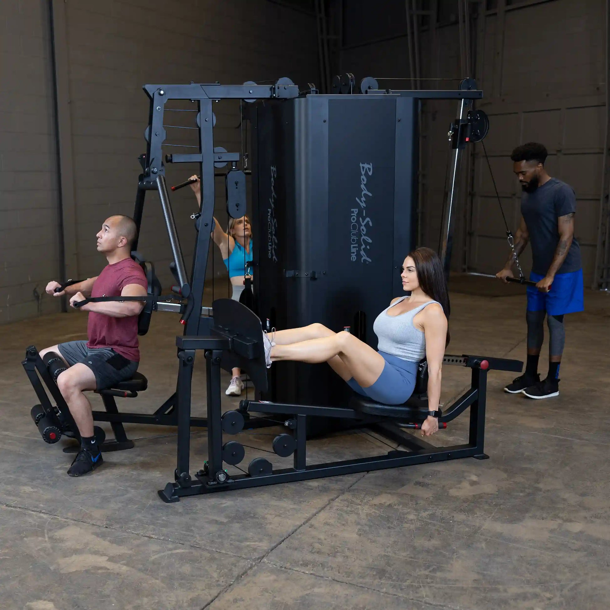 A fitness scene featuring the Body-Solid Pro ClubLine Four-Stack Gym S1000, with individuals using various workout stations, including a seated row, leg press, and cable machine, in a spacious gym environment.