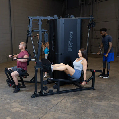 A fitness scene featuring the Body-Solid Pro ClubLine Four-Stack Gym S1000, with individuals using various workout stations, including a seated row, leg press, and cable machine, in a spacious gym environment.