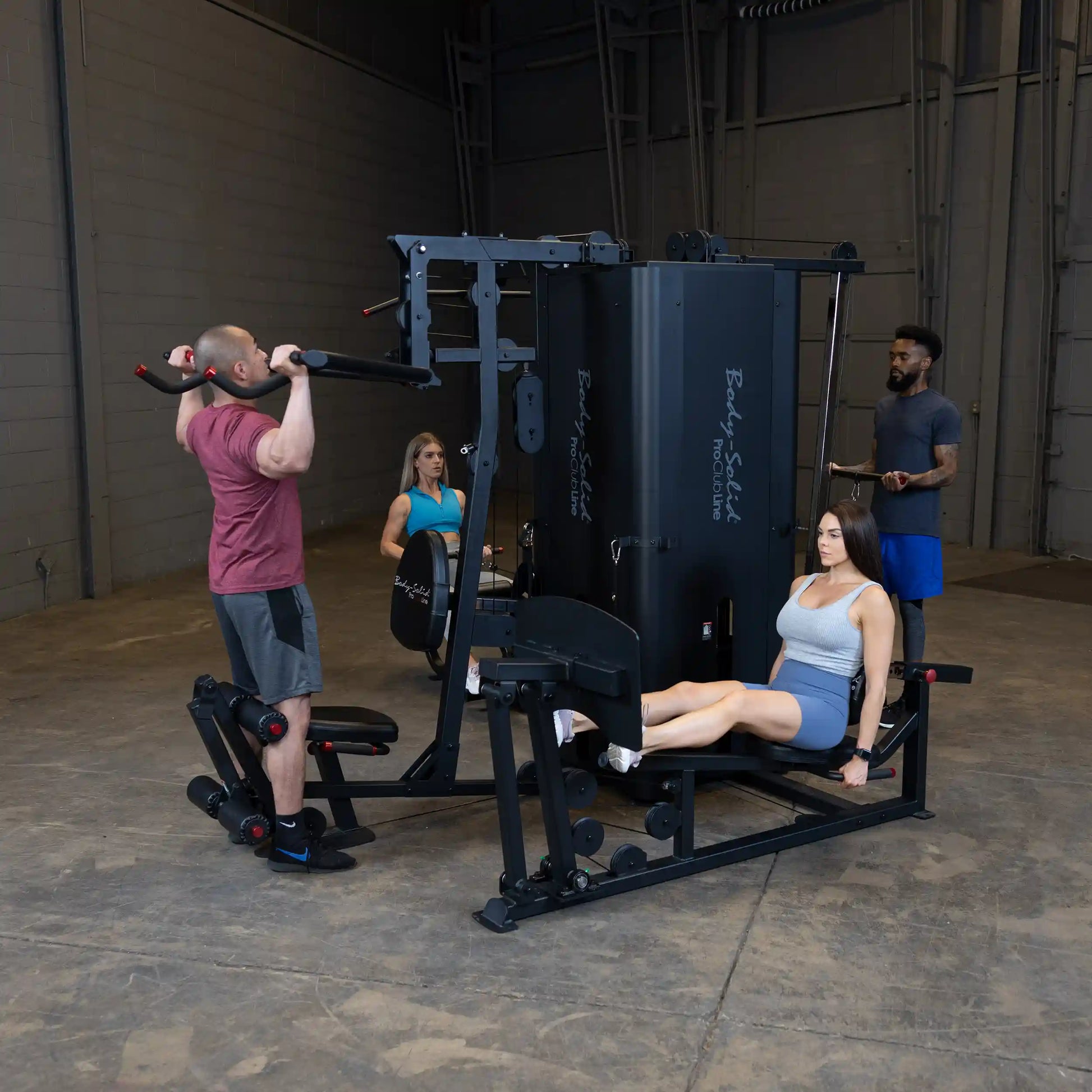 A fitness scene featuring the Body-Solid Pro ClubLine Four-Stack Gym S1000, with a man performing pull-ups, a woman seated on a leg extension machine, and another man instructing in a gym setting.