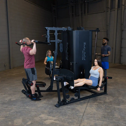 A fitness scene featuring the Body-Solid Pro ClubLine Four-Stack Gym S1000, with a man performing pull-ups, a woman seated on a leg extension machine, and another man instructing in a gym setting.