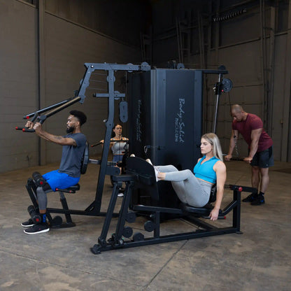 Individuals using the Body-Solid Pro ClubLine Four-Stack Gym S1000, featuring a man performing a seated row, a woman doing leg press, and another person working out in the background.