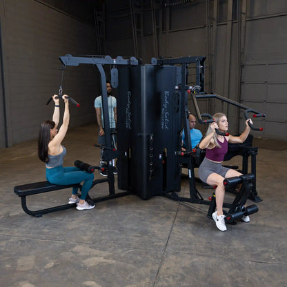 Two women using the Body-Solid Pro ClubLine Four-Stack Gym S1000 for strength training, with a man observing in the background, in a spacious gym setting.
