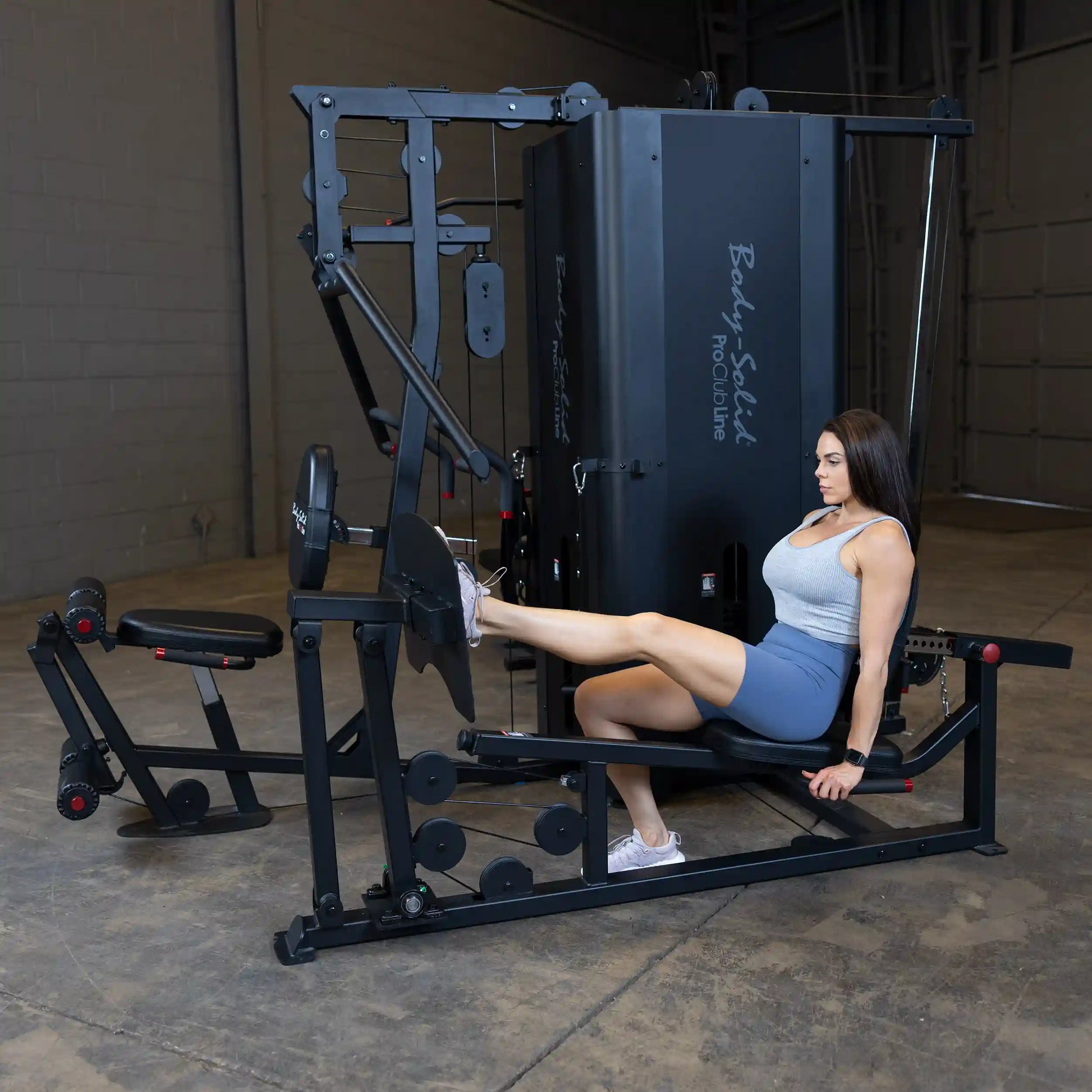 A woman exercises on the Body-Solid Pro ClubLine Four-Stack Gym S1000, using the leg extension attachment in a gym setting.