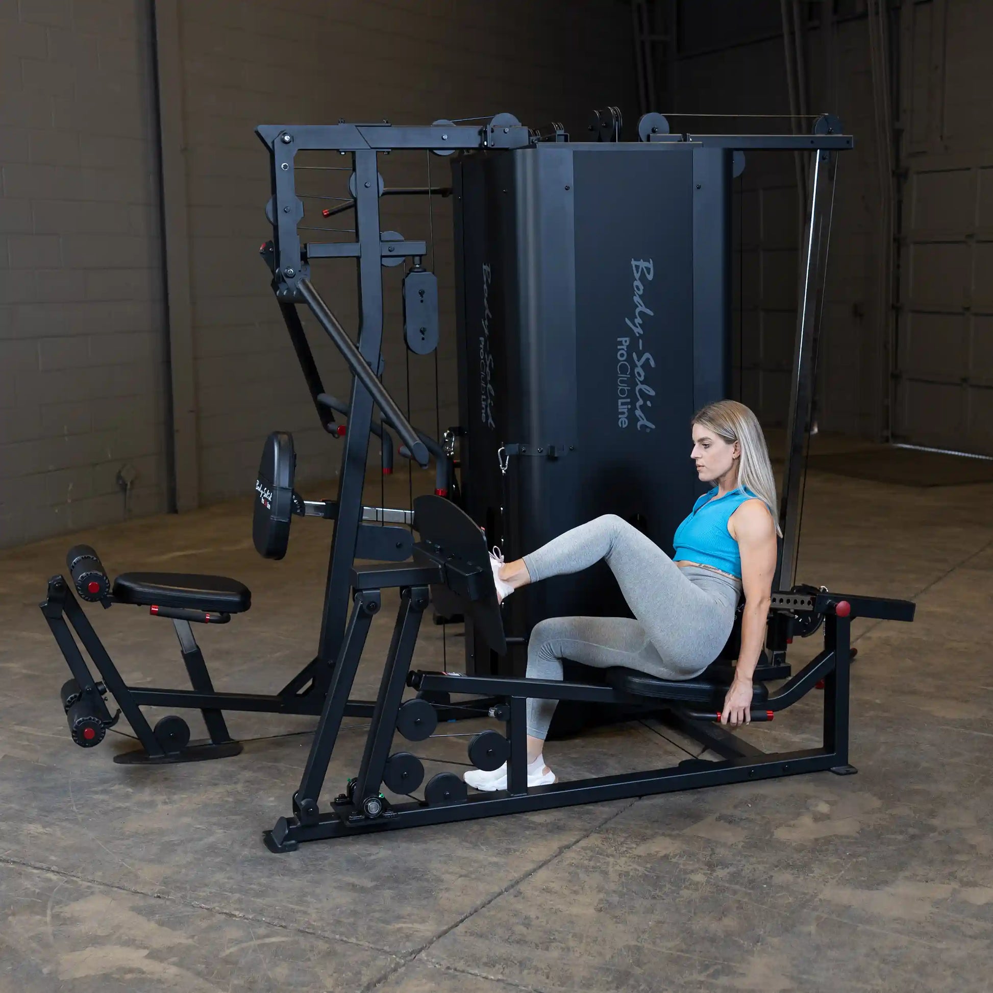 A person using the Body-Solid Pro ClubLine Four-Stack Gym S1000, engaging in a leg exercise while seated on the machine in a spacious gym setting.