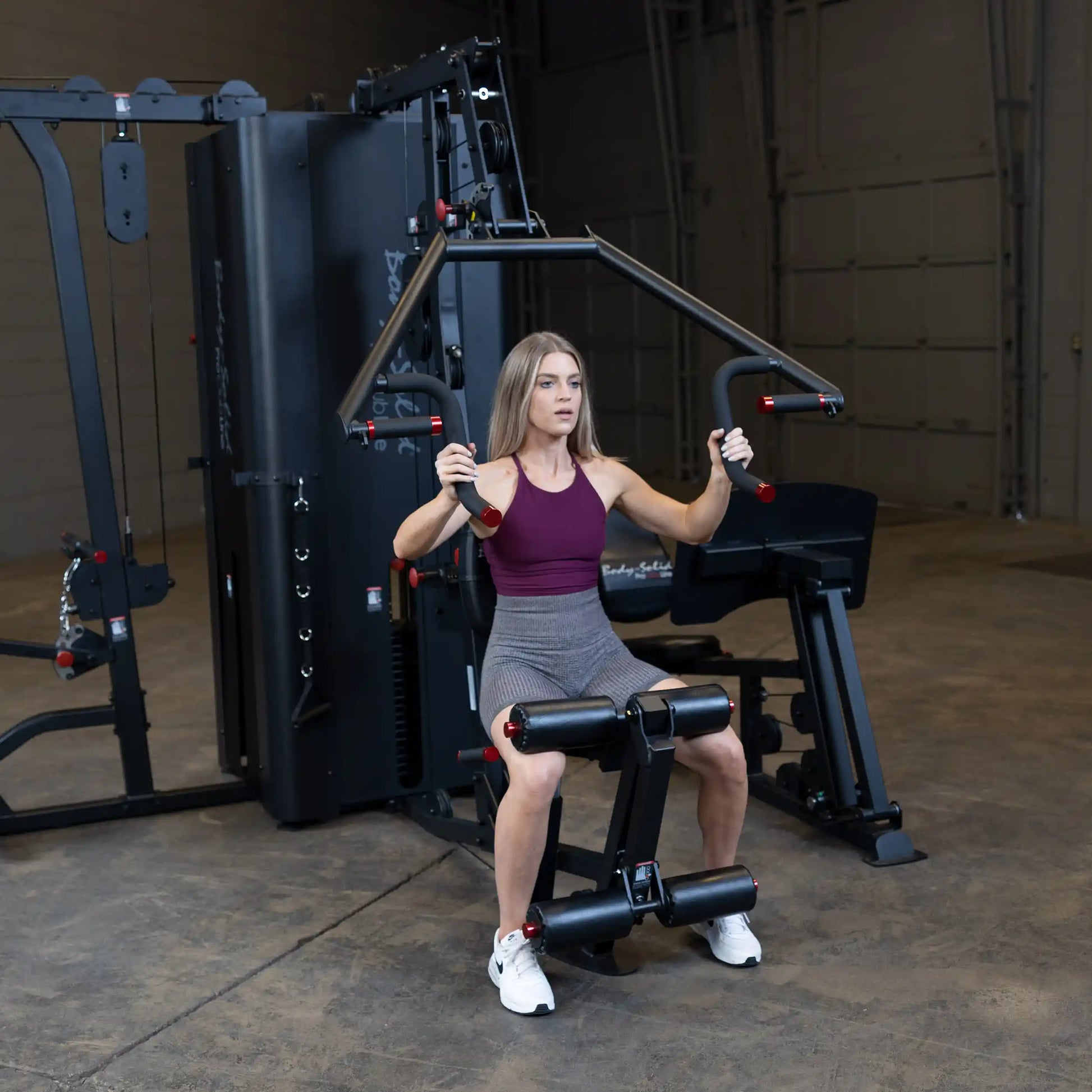 A woman using the Body-Solid Pro ClubLine Four-Stack Gym S1000, performing an exercise while seated on the equipment in a spacious gym setting.