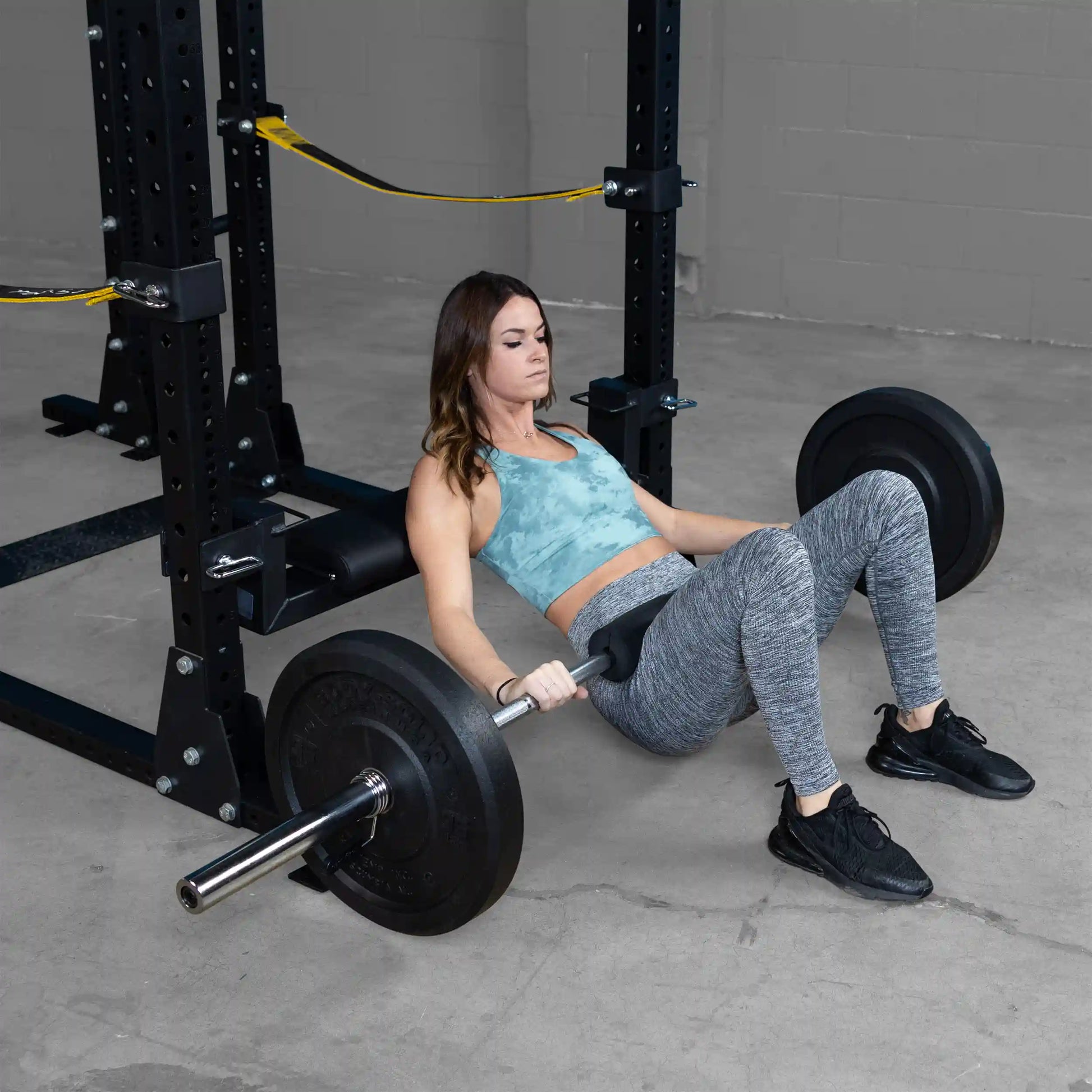 A woman performing hip thrusts using the Body-Solid Pro ClubLine Hip Thrust Attachment SPRHT, with a barbell and weights positioned across her hips, in a gym setting.