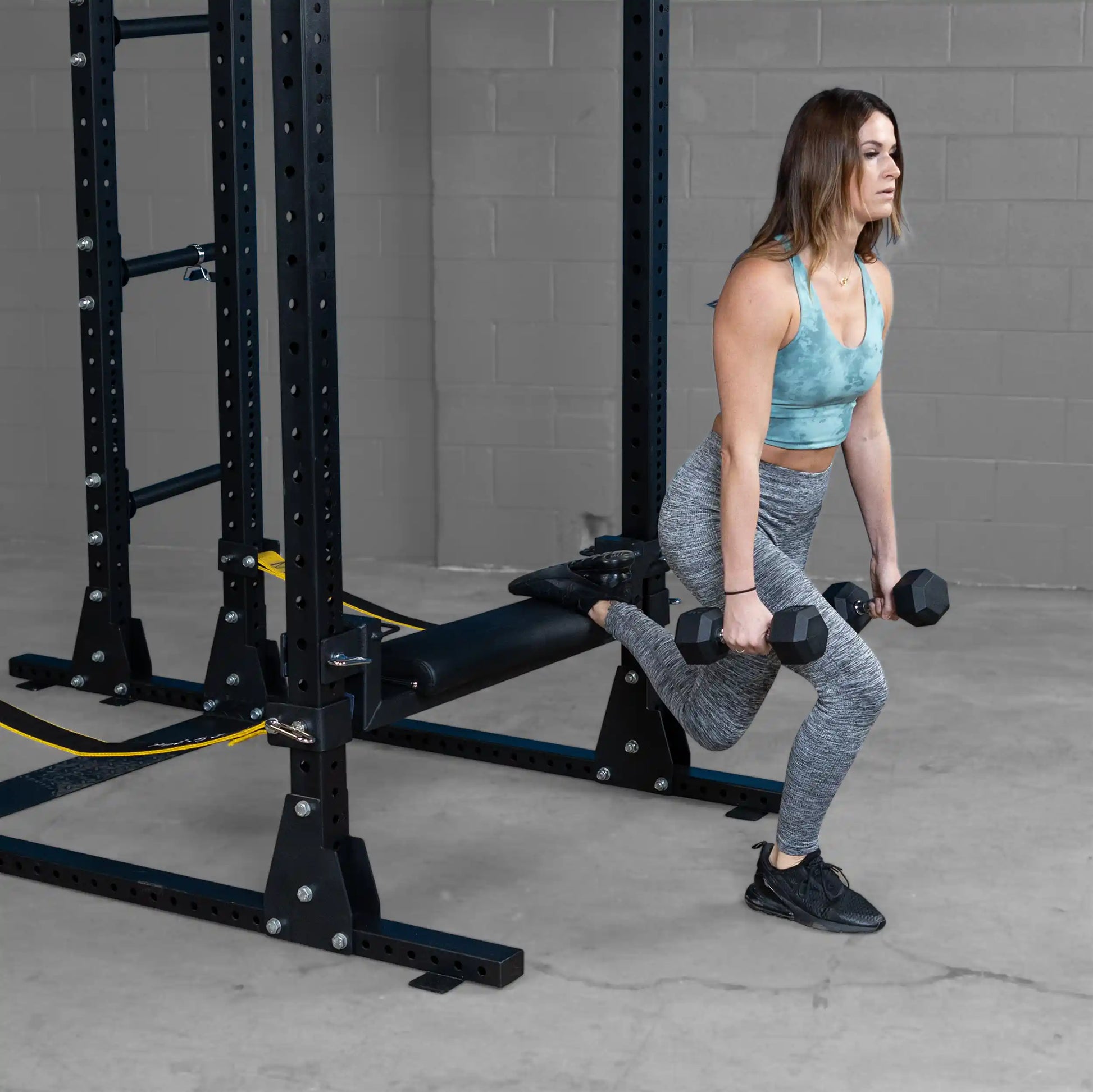 A person performing a single-leg hip thrust using the Body-Solid Pro ClubLine Hip Thrust Attachment SPRHT in a gym setting, demonstrating proper exercise form with dumbbells in hand.