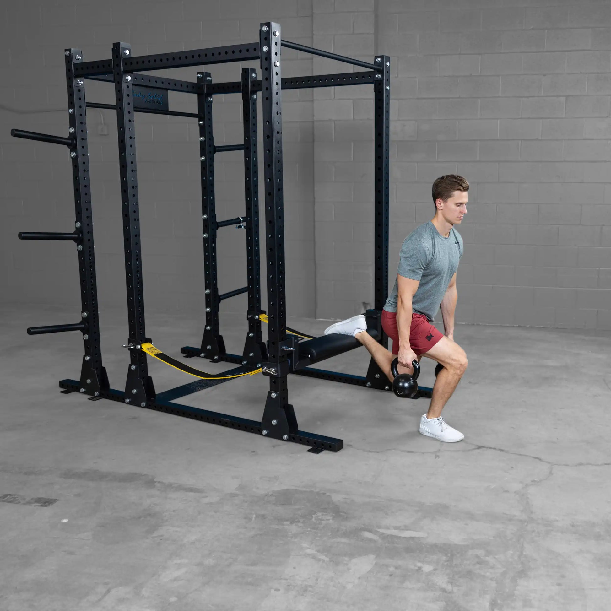 A person demonstrating an exercise using the Body-Solid Pro ClubLine Hip Thrust Attachment SPRHT, positioned in front of a squat rack, while holding a kettlebell in one hand.