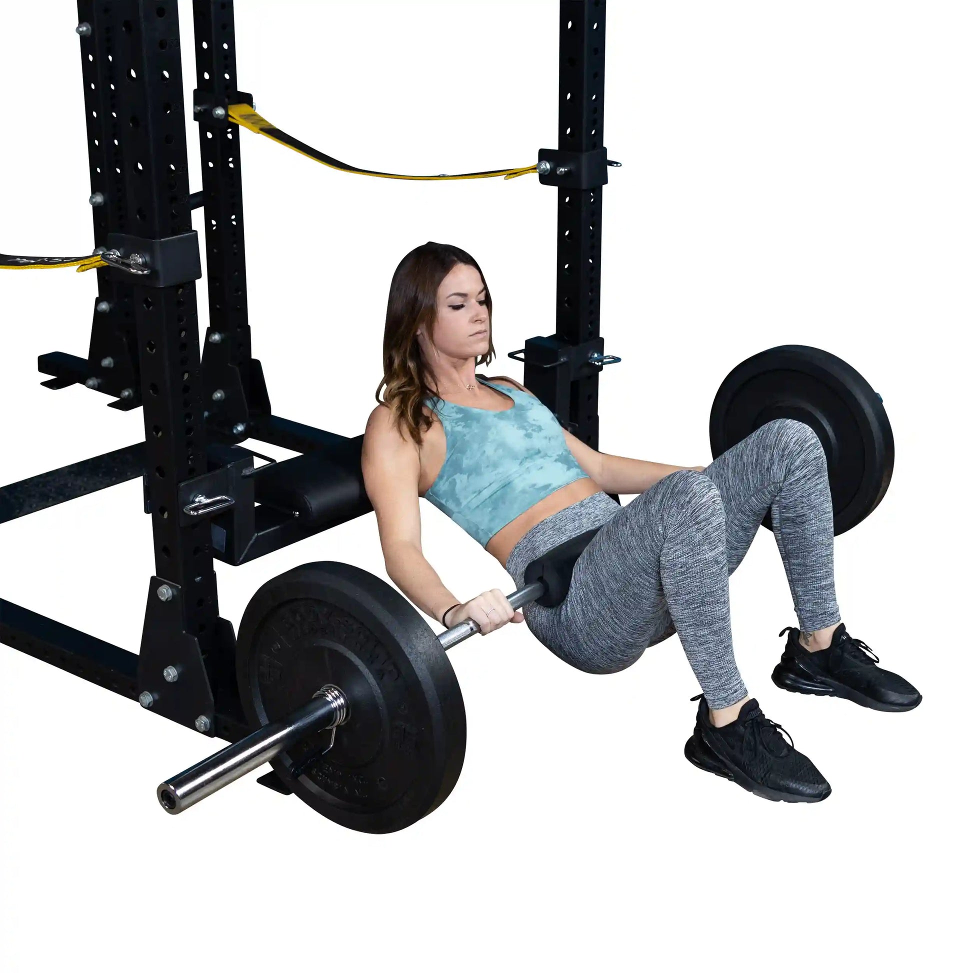 A woman performing hip thrusts using the Body-Solid Pro ClubLine Hip Thrust Attachment SPRHT, with a barbell loaded with weight plates, positioned on a squat rack. She is wearing a blue athletic top and gray leggings.