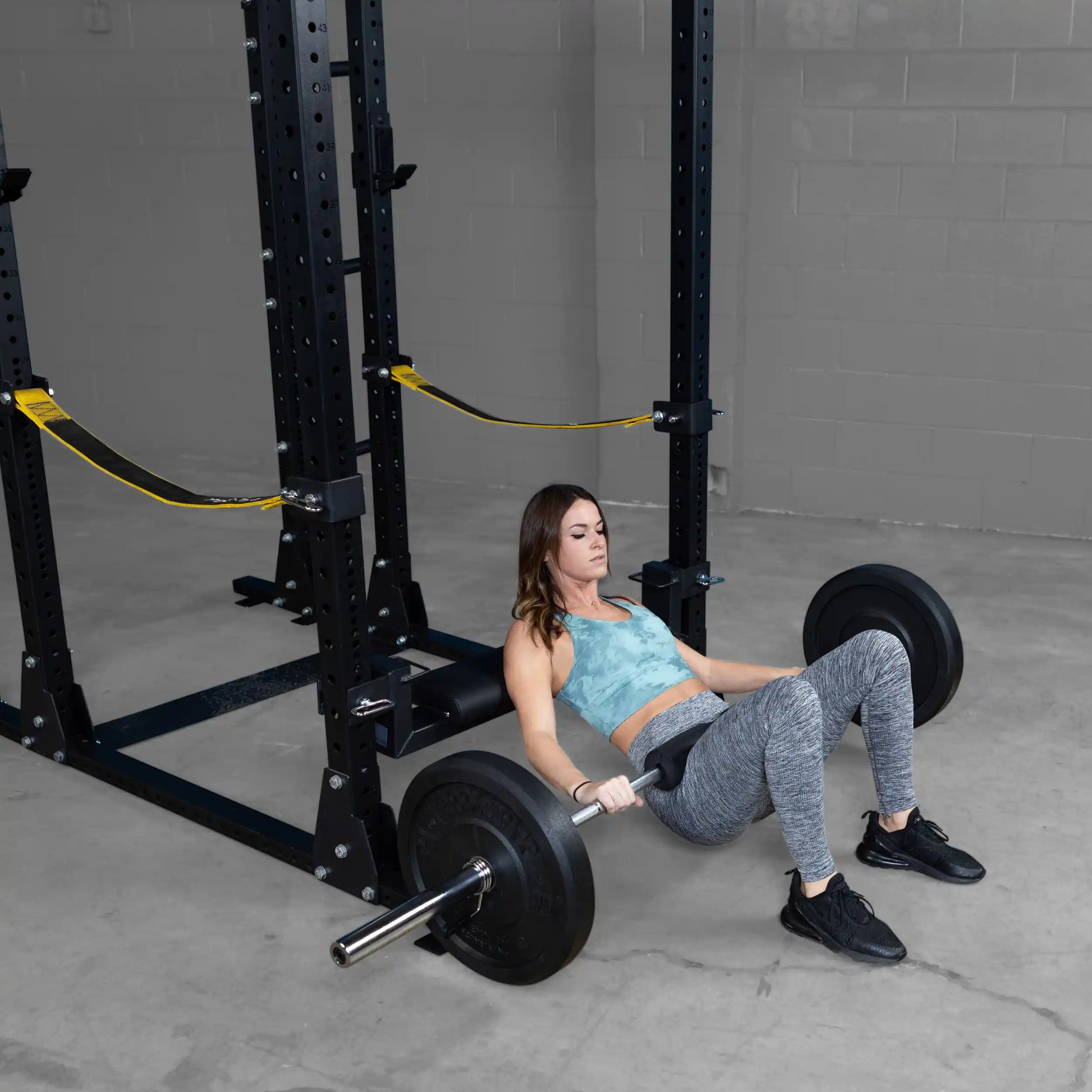 Woman performing a hip thrust exercise using the Body-Solid Pro ClubLine Hip Thrust Attachment SPRHT, with a weight bar and squat rack.