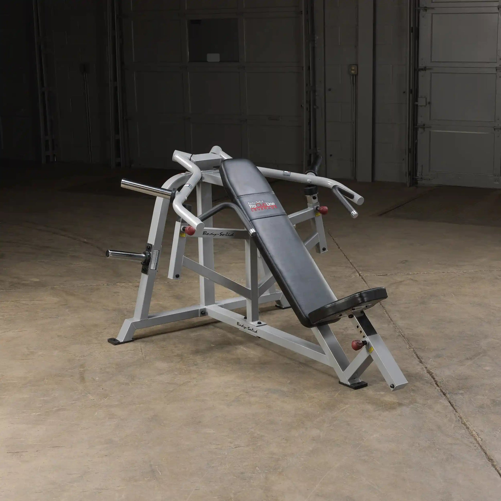 Body-Solid Pro ClubLine Leverage Incline Press LVIP, a strength training machine with a padded seat and adjustable arms, positioned on a concrete floor in a dimly lit space.