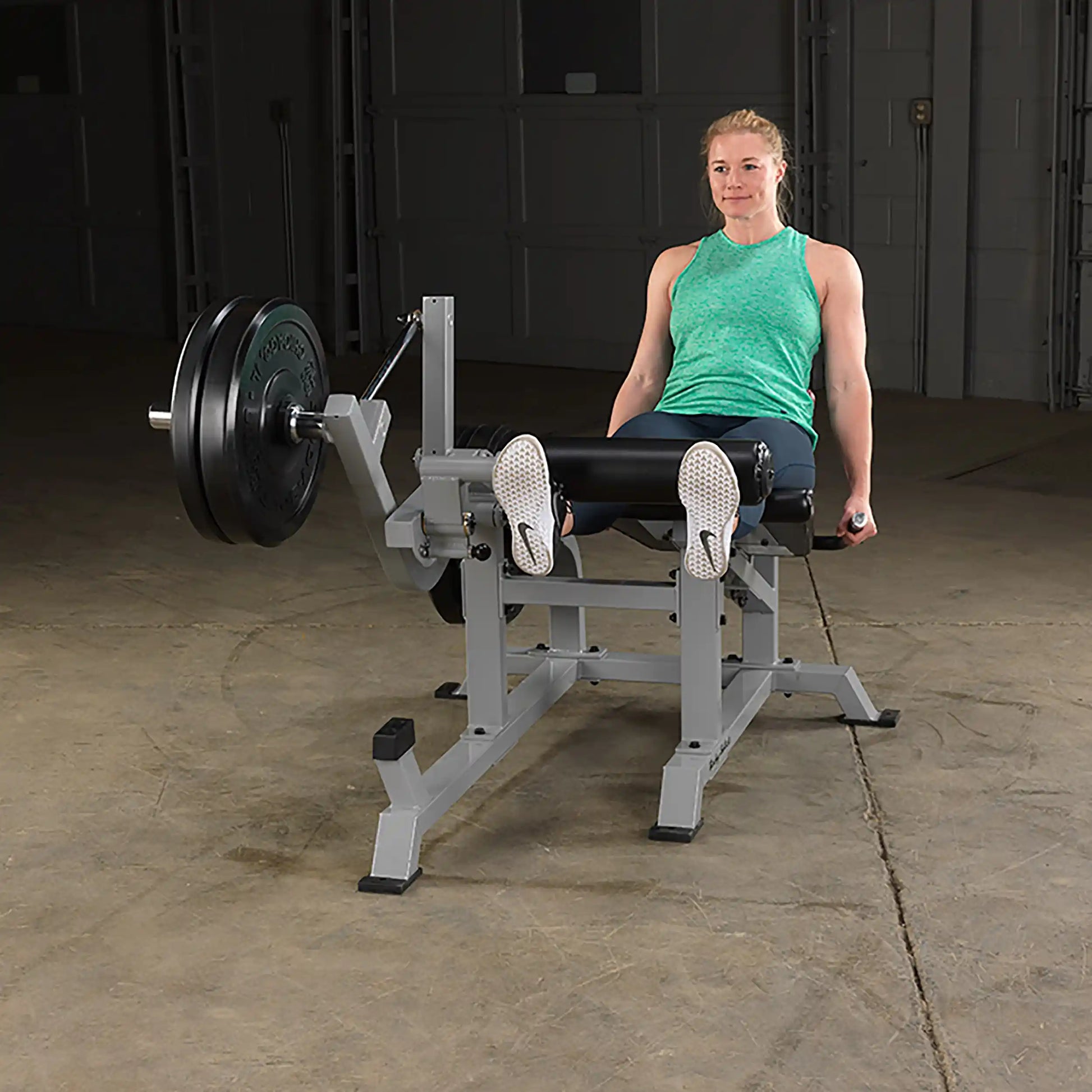 A woman demonstrating the Body-Solid Pro ClubLine Leverage Leg Extension Machine LVLE, seated with her legs extended, in a gym setting.