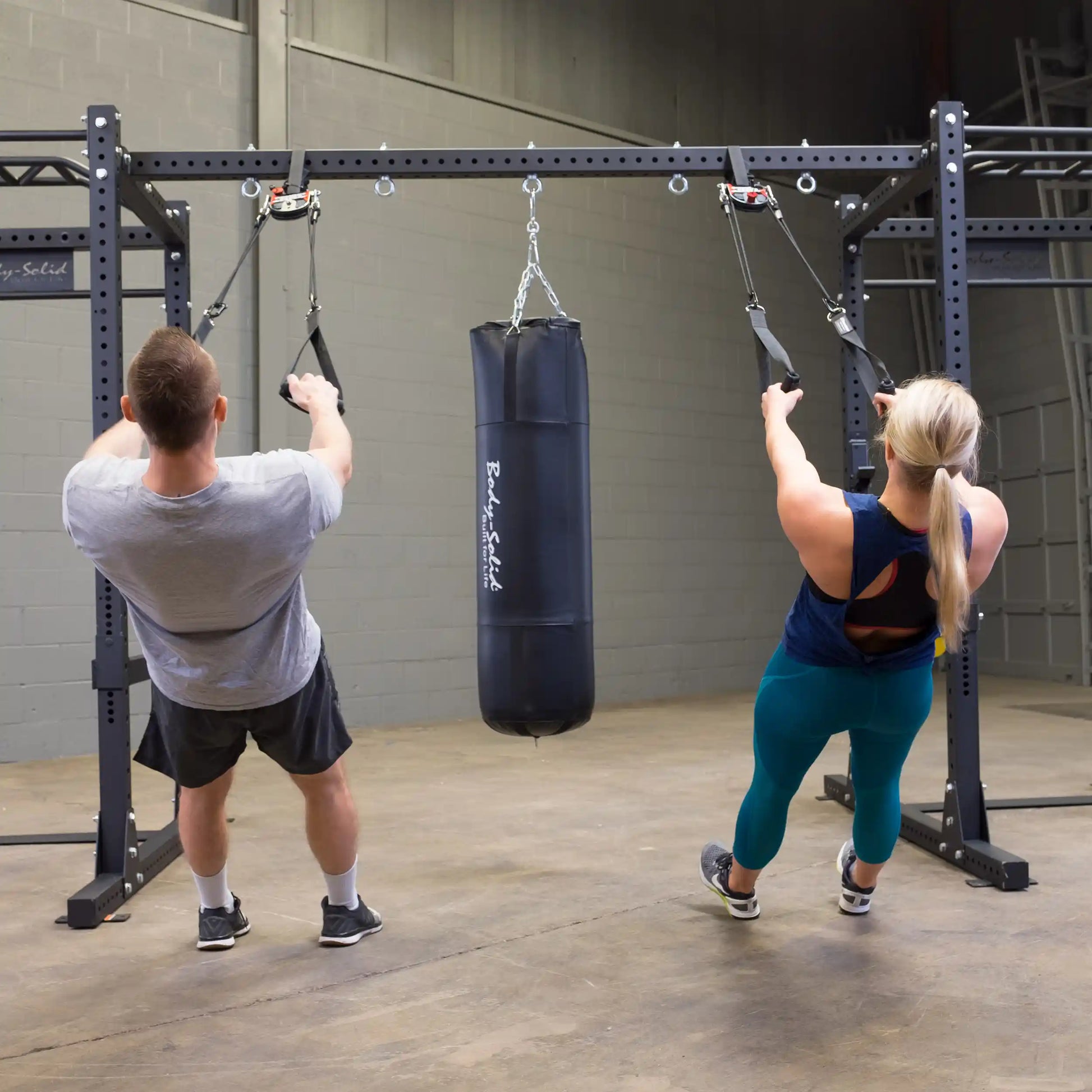 Two people exercising with resistance straps attached to a Body-Solid Pro ClubLine Power Rack Connecting Bar SPRACB, with a punching bag hanging in the center.