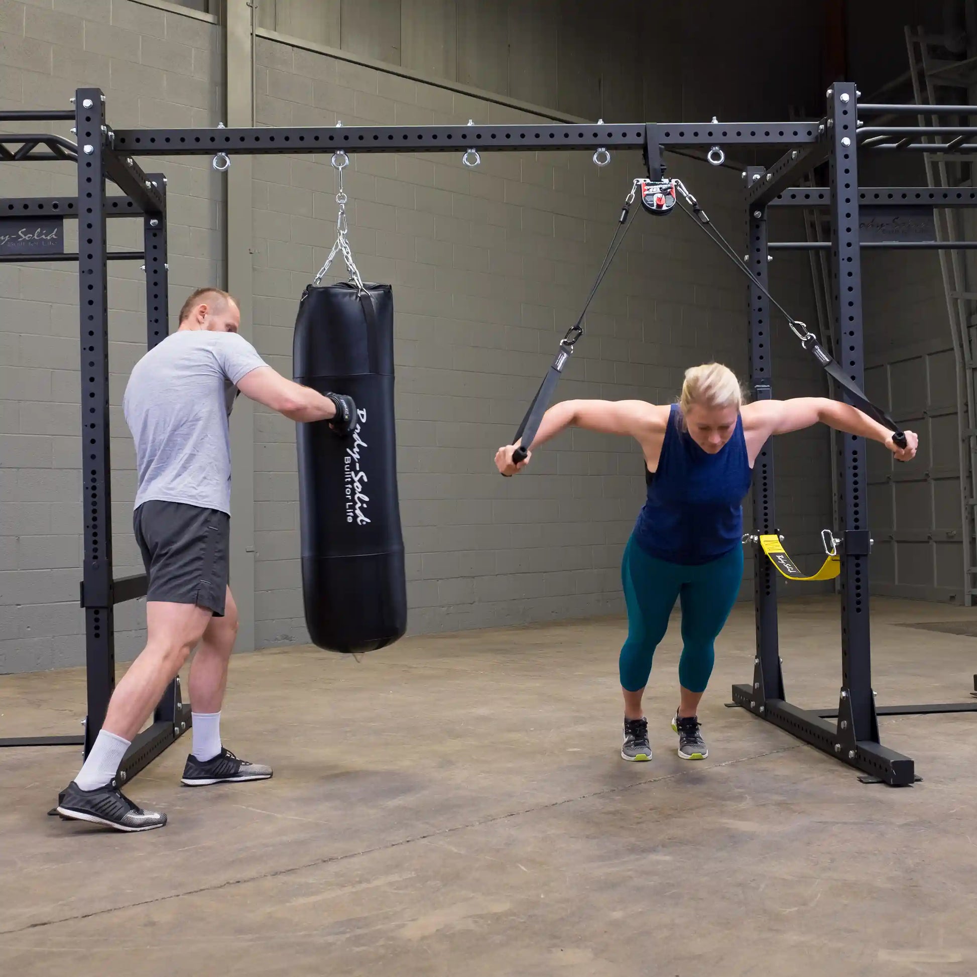 A woman exercises with resistance cables attached to a Body-Solid Pro ClubLine Power Rack Connecting Bar SPRACB, while a man punches a hanging punching bag in a gym setting.