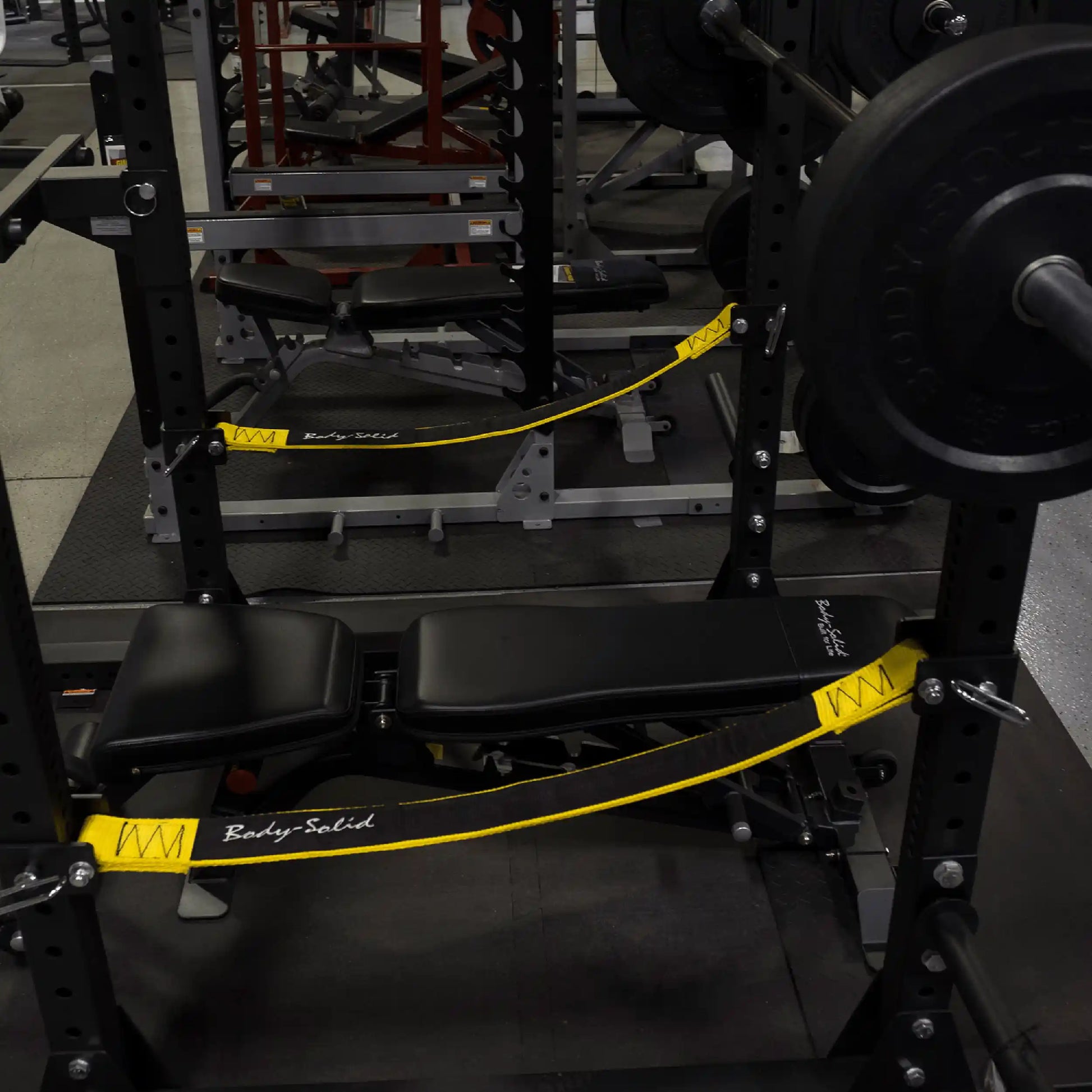 Close-up of Body-Solid Pro ClubLine Power Rack Safety Straps SPRSS in a gym setting, featuring a black bench and weight plates in the background.