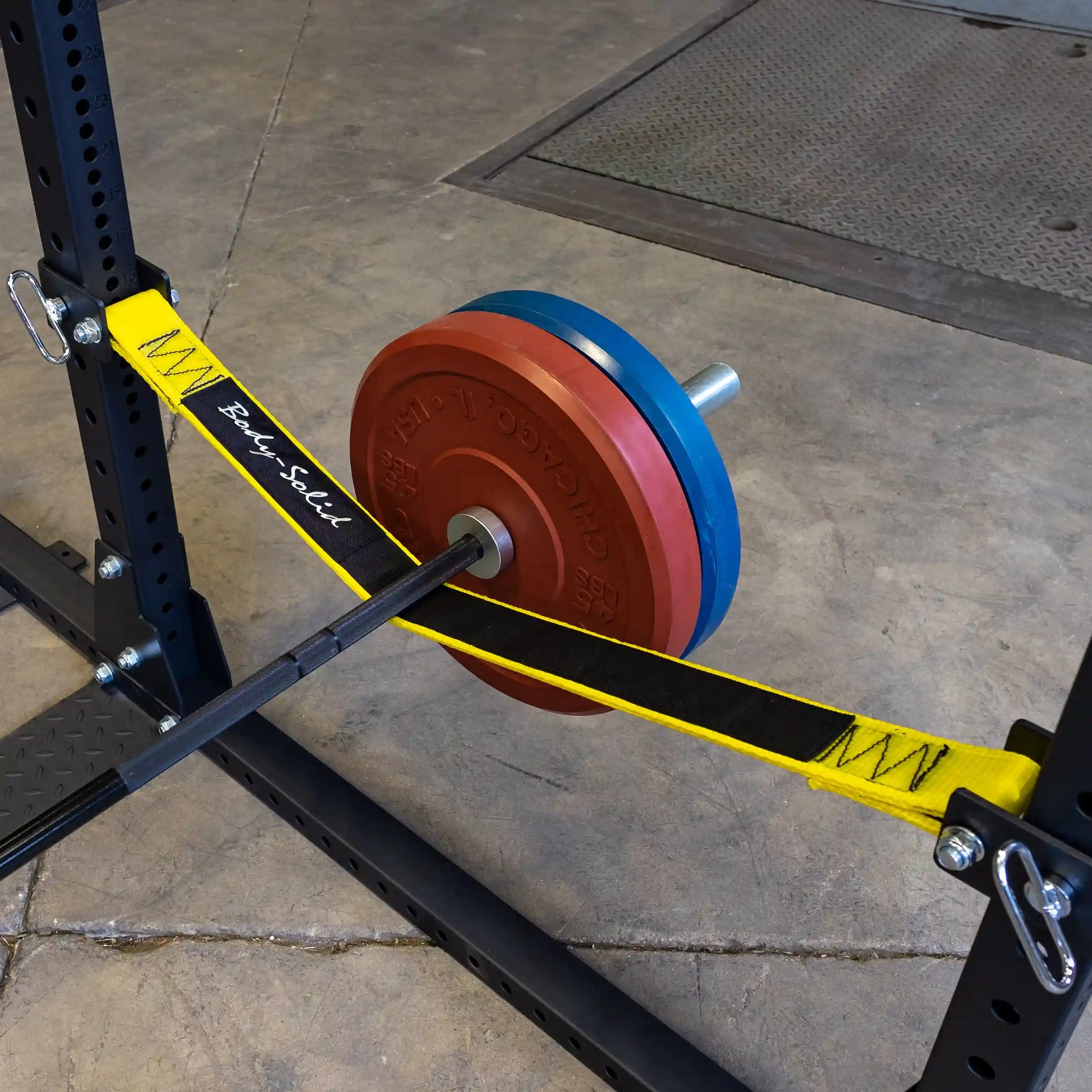A close-up view of the Body-Solid Pro ClubLine Power Rack Safety Straps SPRSS, featuring a barbell with colored weight plates secured in place by the yellow and black safety straps. The background shows a gym floor with a textured surface.