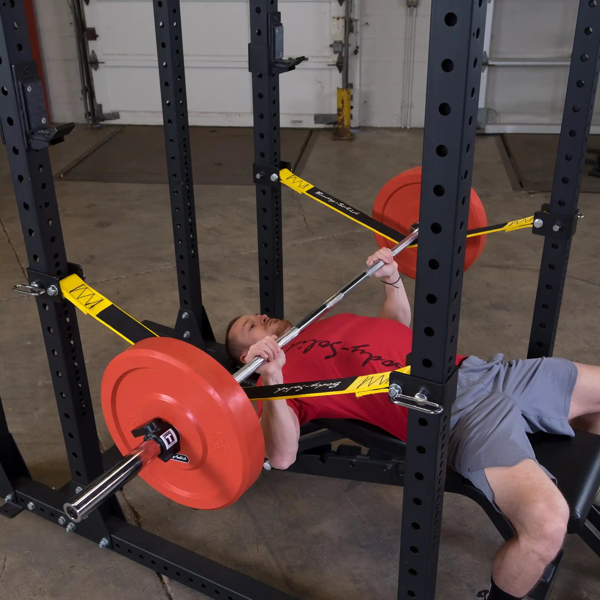 A person performing a bench press using the Body-Solid Pro ClubLine Power Rack Safety Straps SPRSS, with a barbell and weights on the rack in a gym setting.