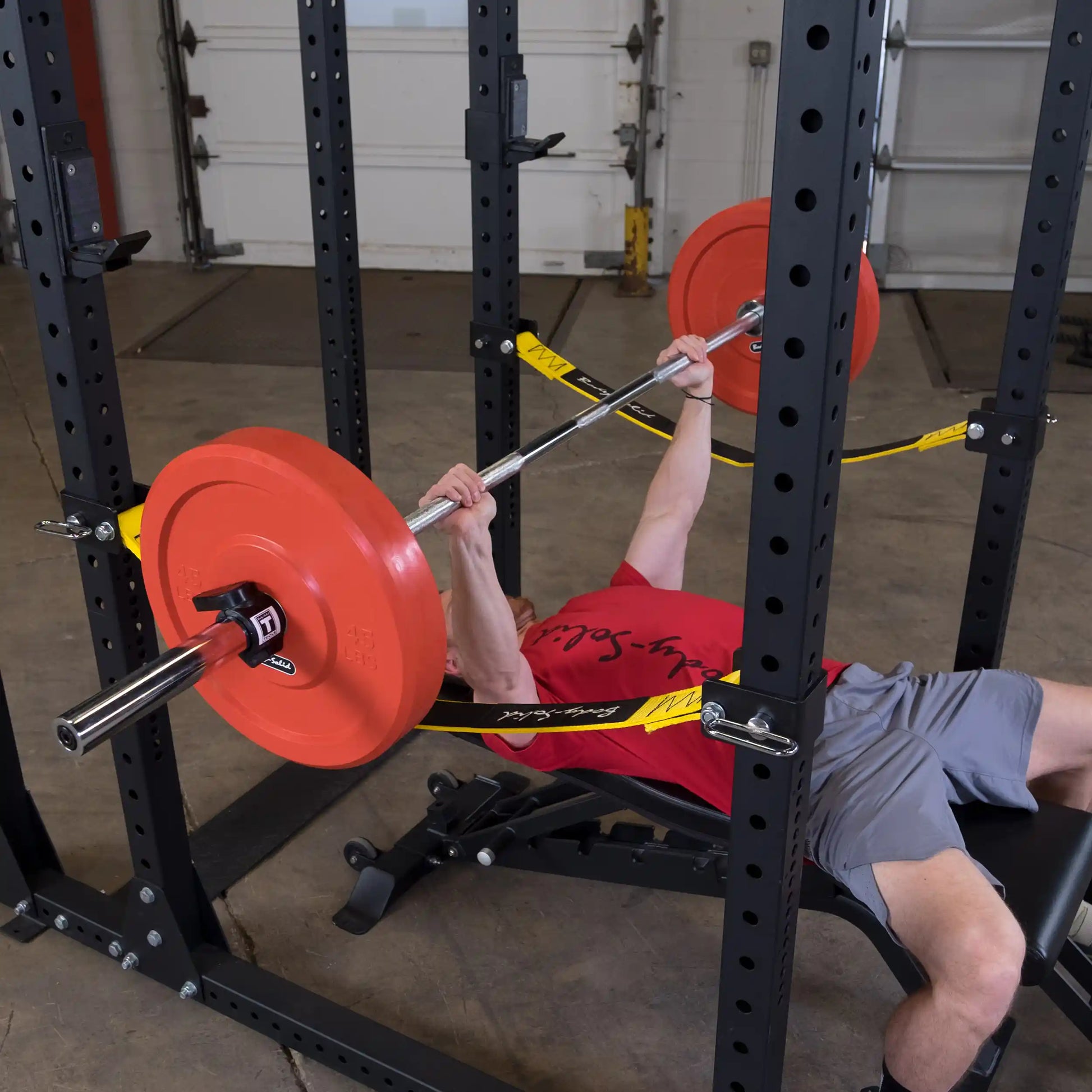 A person performing a bench press with a barbell and large weights, using the Body-Solid Pro ClubLine Power Rack Safety Straps SPRSS for added safety in a gym setting.