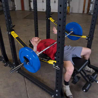 A person performing a bench press in a gym using the Body-Solid Pro ClubLine Power Rack Safety Straps SPRSS, featuring blue weight plates and a black bench.