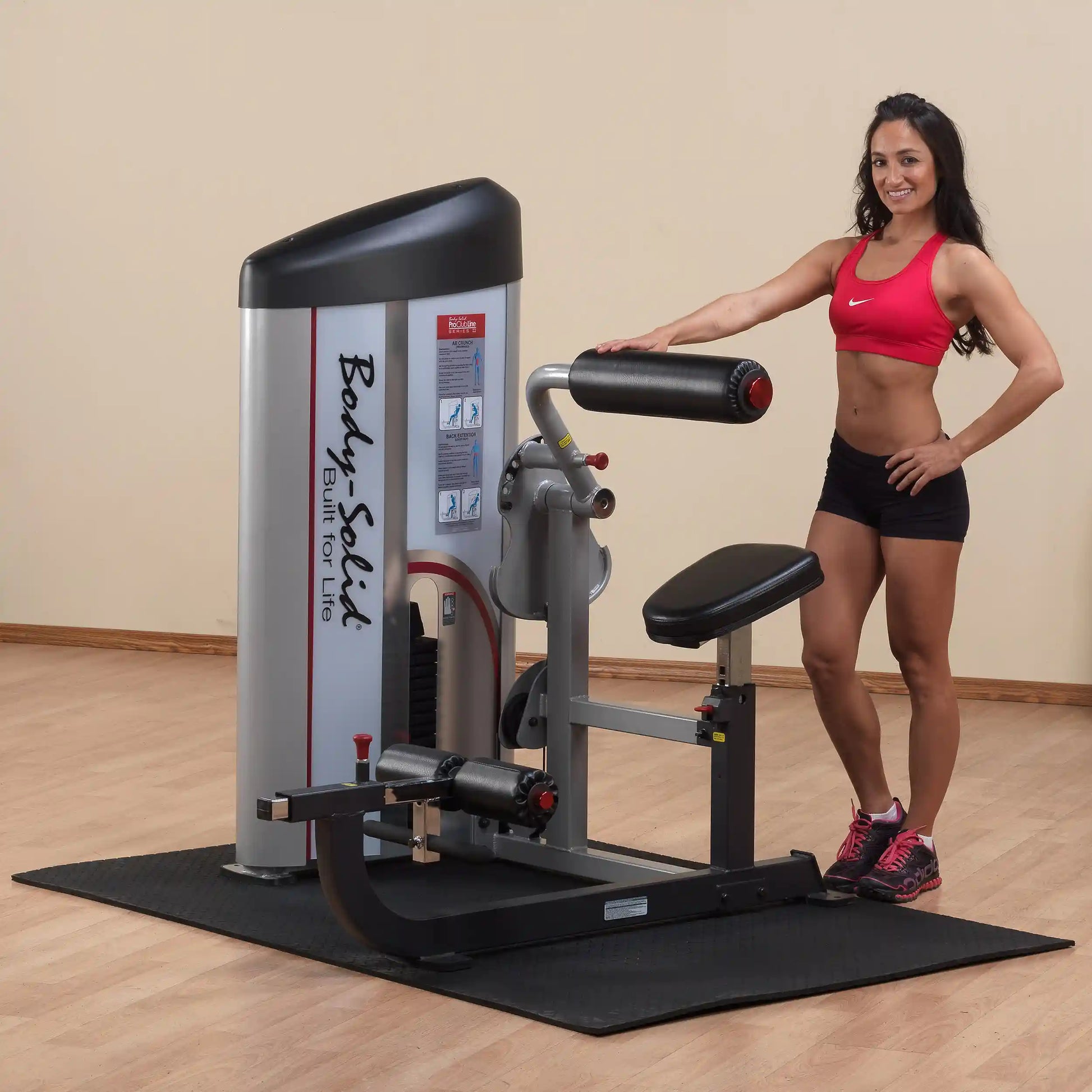 A fitness model stands next to the Body-Solid Pro ClubLine Series 2 Ab & Back Machine S2ABB in a gym setting, showcasing the equipment designed for abdominal and back workouts.