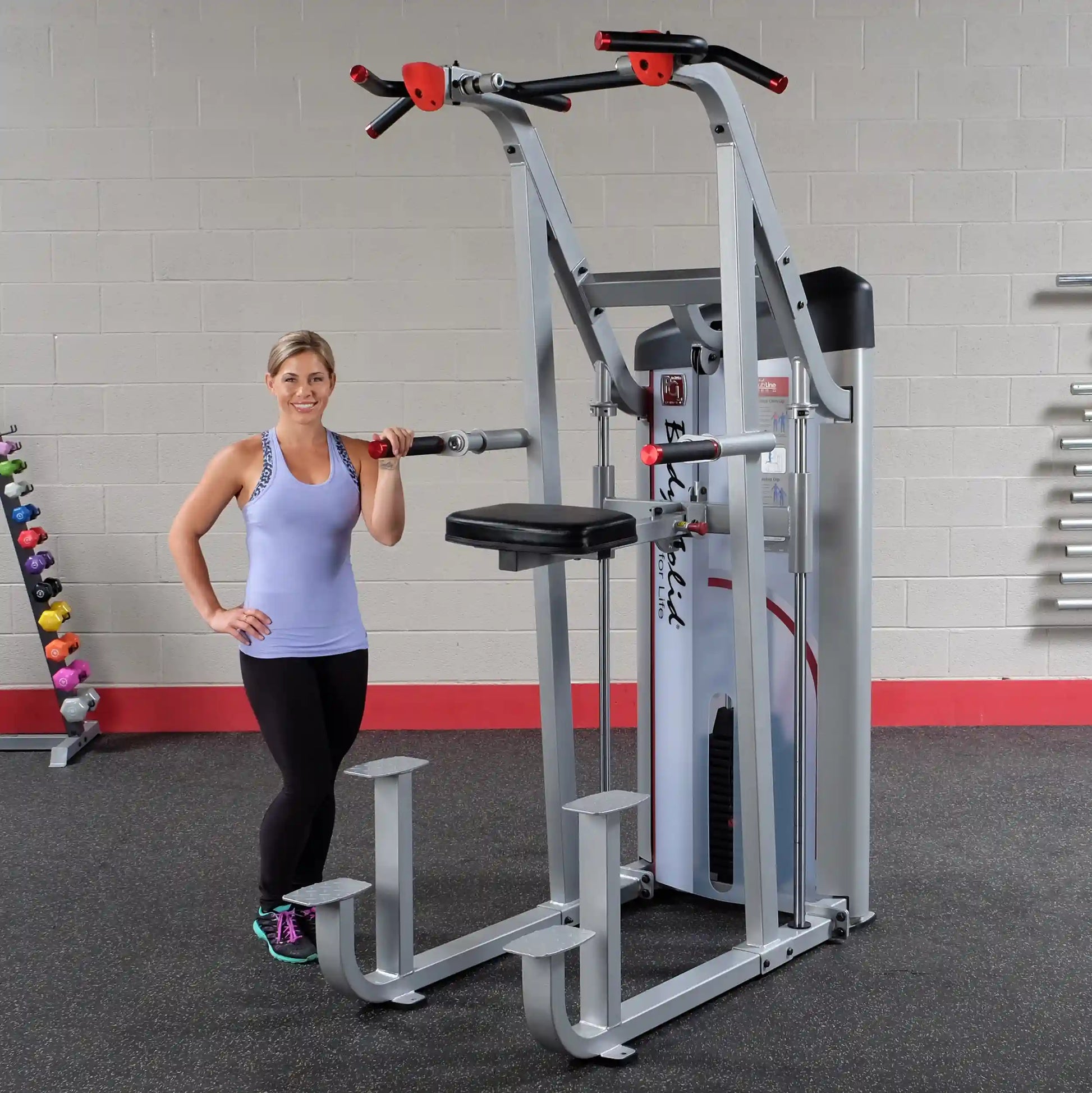 A woman stands next to the Body-Solid Pro ClubLine Series 2 Assisted Chin & Dip Machine S2ACD in a gym setting, showcasing the equipment designed for assisted chin-ups and dips.