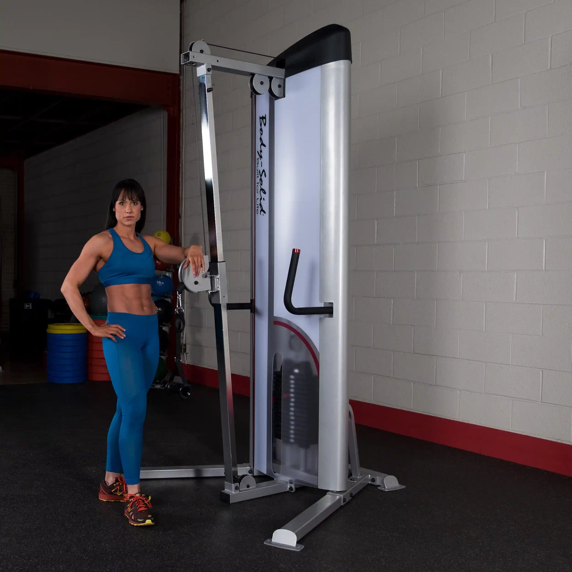 A fit woman in a blue athletic outfit poses next to the Body-Solid Pro ClubLine Series 2 Cable Column S2CC in a gym setting.