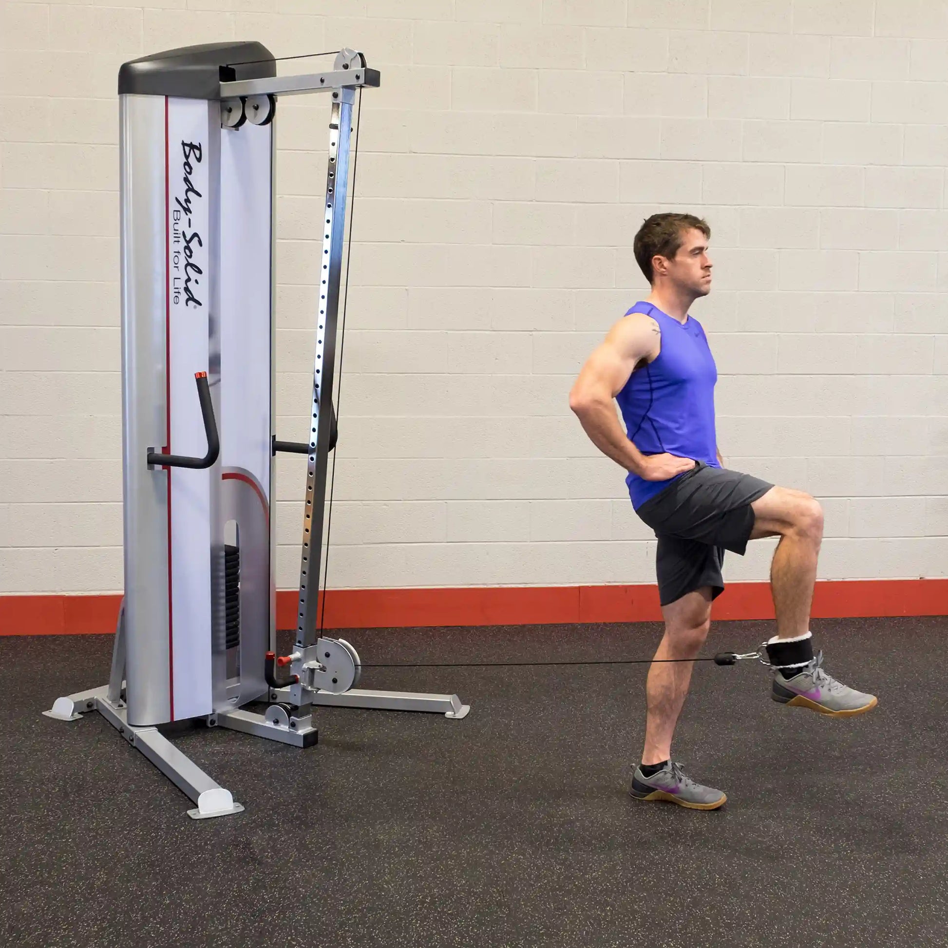 A man in a blue tank top and shorts performs an exercise using the Body-Solid Pro ClubLine Series 2 Cable Column S2CC, with one leg raised and a cable attachment around his ankle. The gym setting features a light-colored wall and a dark floor.