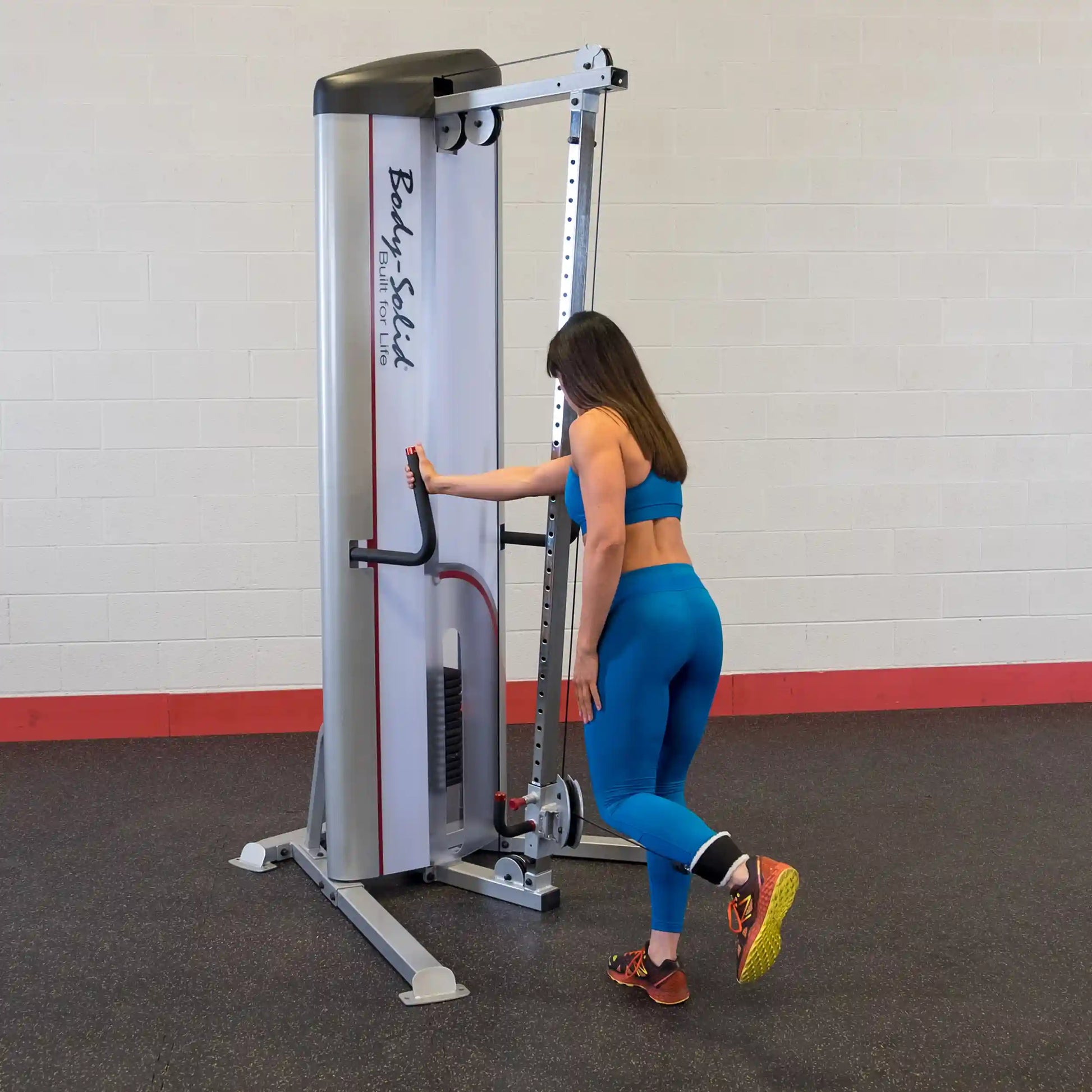 A woman in blue workout attire is using the Body-Solid Pro ClubLine Series 2 Cable Column S2CC for exercise, standing beside the machine with one leg raised behind her.