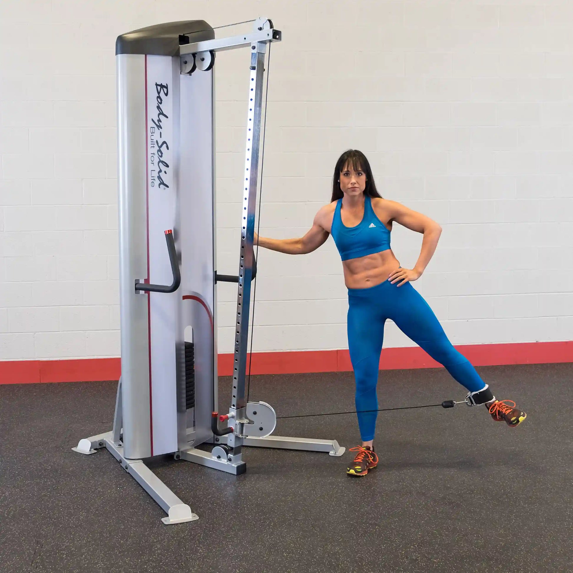 A woman in a blue sports bra and leggings demonstrates an exercise using the Body-Solid Pro ClubLine Series 2 Cable Column S2CC, standing beside the equipment with one leg extended and holding onto the machine for support.