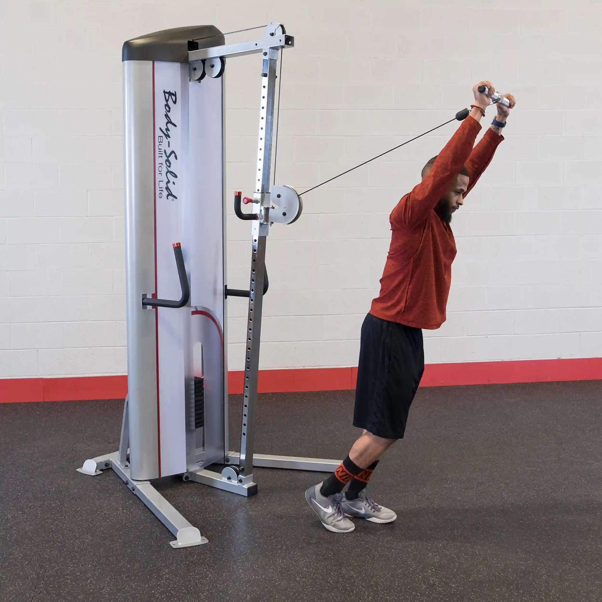 A person performing an exercise using the Body-Solid Pro ClubLine Series 2 Cable Column S2CC, standing with arms extended overhead while gripping the cable handles. The setting features a gym interior with a textured floor and a light-colored wall.