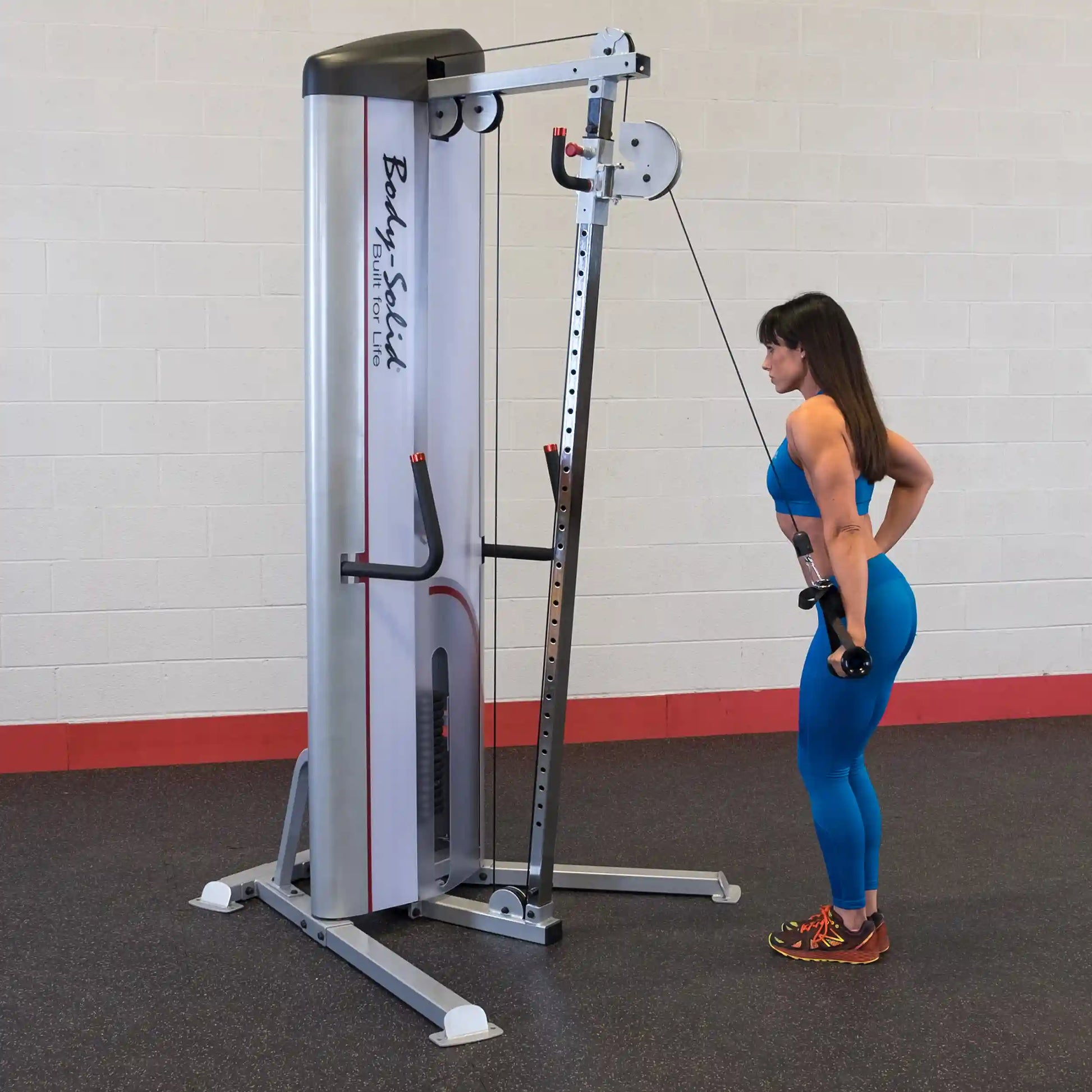 A woman in a blue workout outfit is performing an exercise with dumbbells in front of the Body-Solid Pro ClubLine Series 2 Cable Column S2CC, set in a gym with a light-colored wall and a red accent.